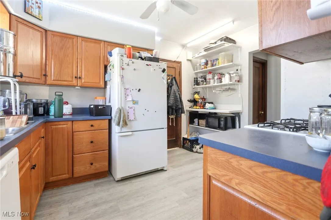 Kitchen featuring white appliances, brown cabinetry, light wood-style floors, dark countertops, and a ceiling fan
