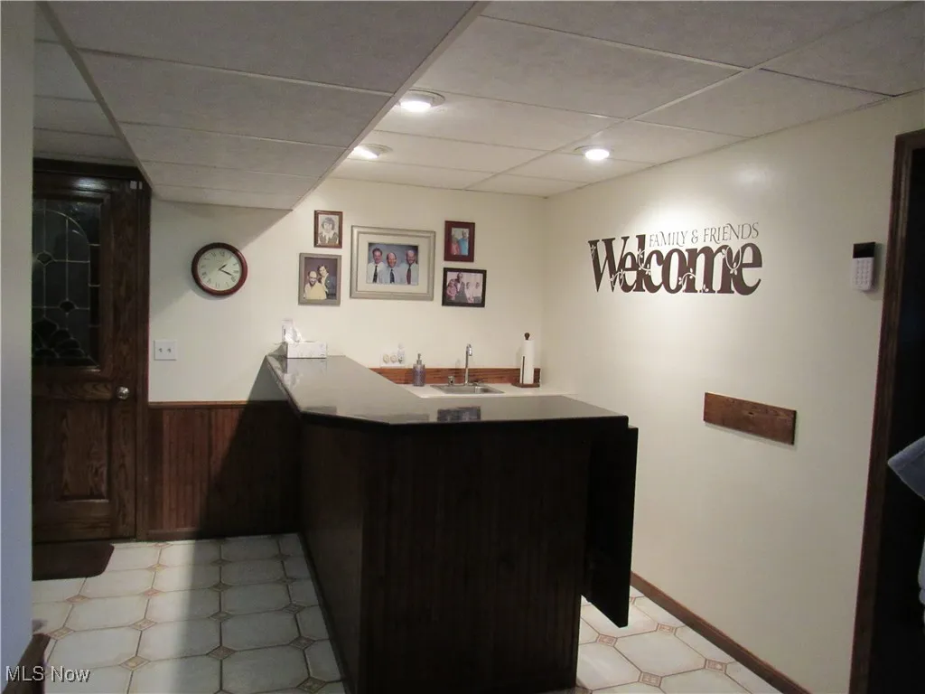 Indoor wet bar featuring a drop ceiling, wainscoting, dark countertops, sink and dark brown cabinets