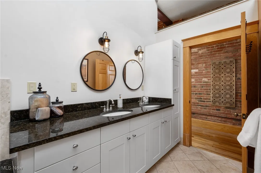 Bathroom featuring double vanity, light tile patterned flooring, and brick wall