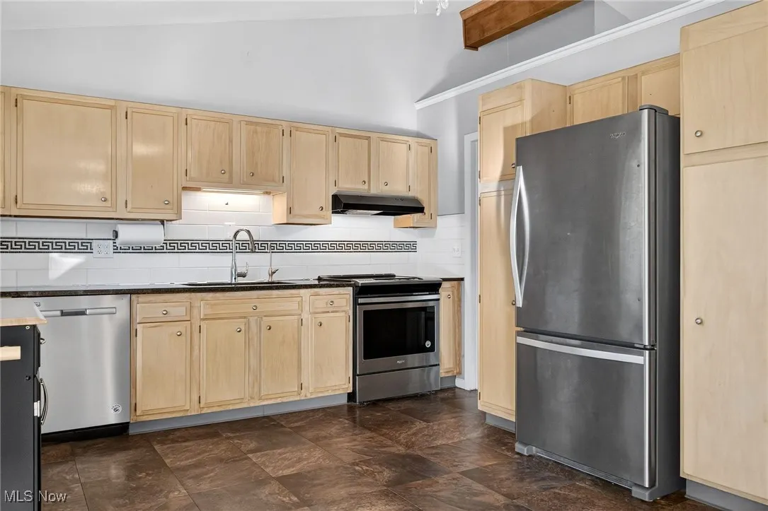 Kitchen with light brown cabinetry, stainless steel appliances, tasteful backsplash, dark stone countertops, and lofted ceiling
