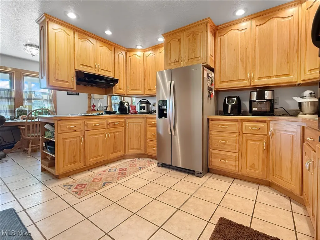 Kitchen with stainless steel fridge, light tile patterned flooring, a textured ceiling, under cabinet range hood, and a peninsula