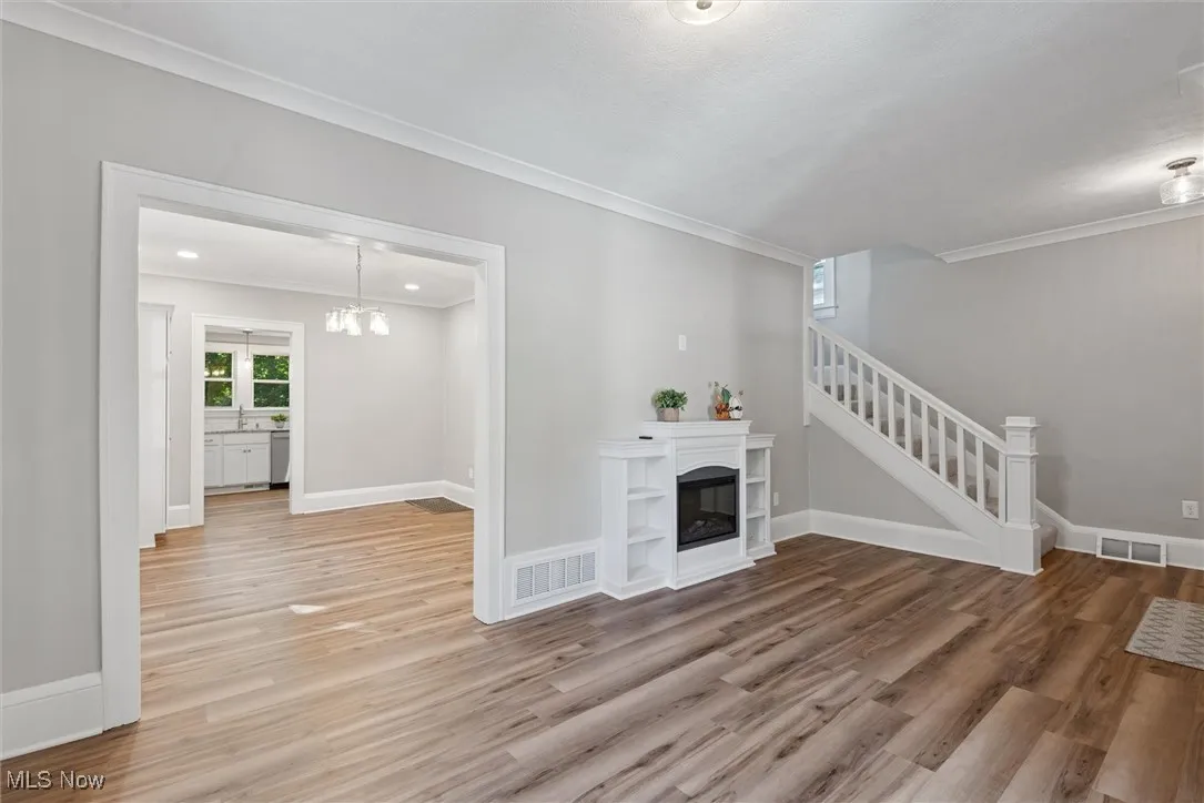Unfurnished living room with a glass covered fireplace, light wood-type flooring, ornamental molding, a chandelier, and stairway