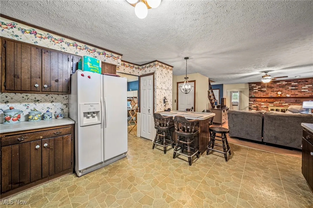 Kitchen with dark brown cabinets, white fridge with ice dispenser, light countertops, light stone finish flooring, and ceiling fan