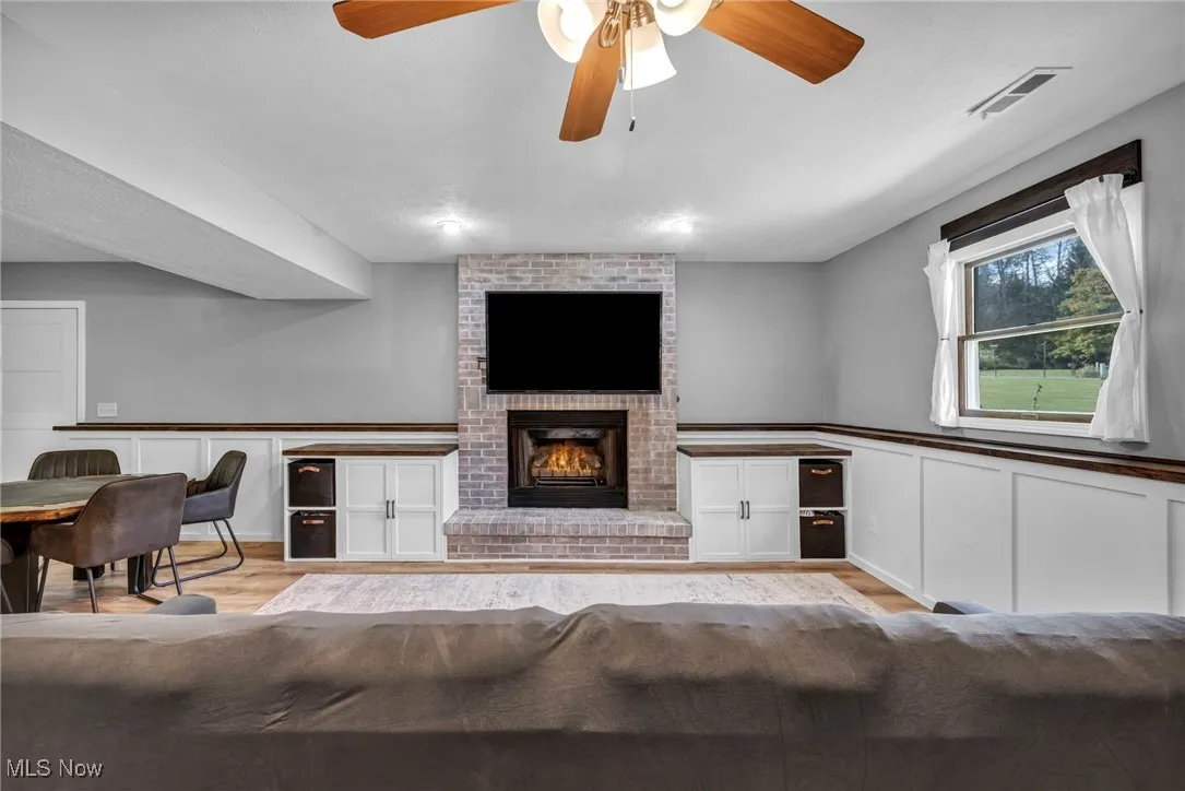 Living area with a brick fireplace, a decorative wall, ceiling fan, wood finished floors, and a wainscoted wall