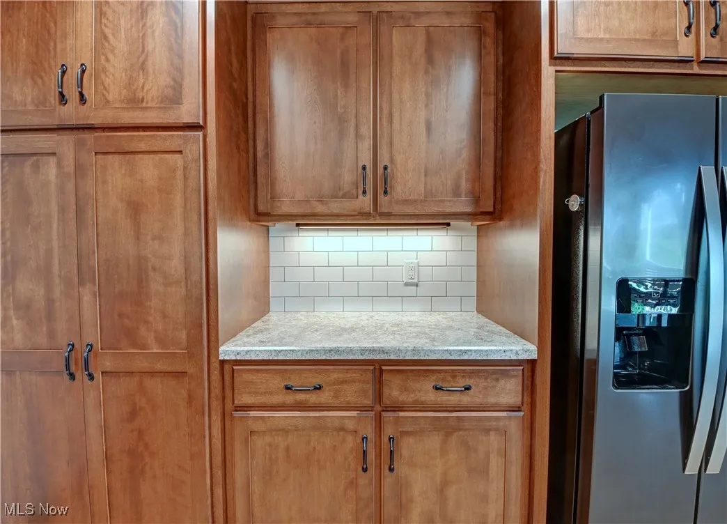 Kitchen with stainless steel refrigerator with ice dispenser, tasteful backsplash, brown cabinetry, and light stone counters