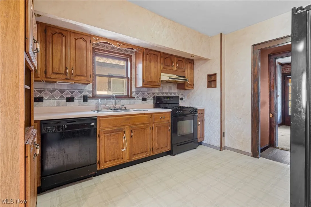 Kitchen with brown cabinetry, black appliances, light countertops, tasteful backsplash, and under cabinet range hood