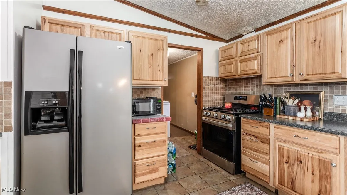 Kitchen with appliances with stainless steel finishes, a textured ceiling, light brown cabinets, vaulted ceiling, and light tile patterned floors
