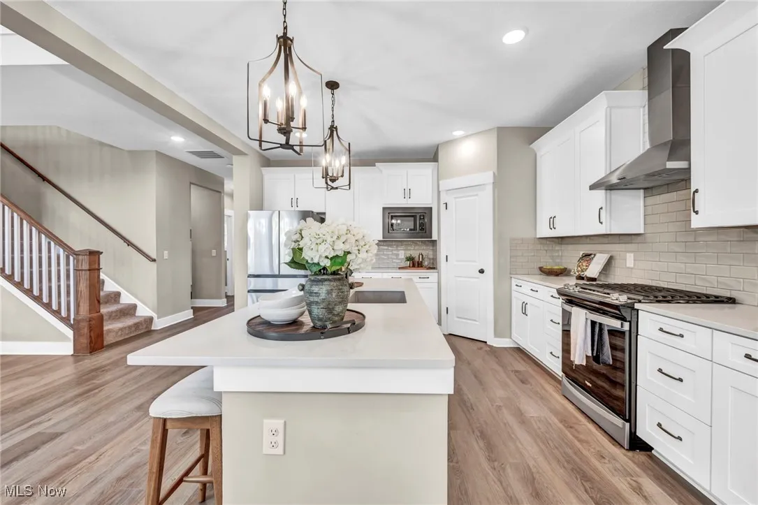 Kitchen featuring stainless steel appliances, decorative light fixtures, an island with sink, wall chimney exhaust hood, and white cabinetry