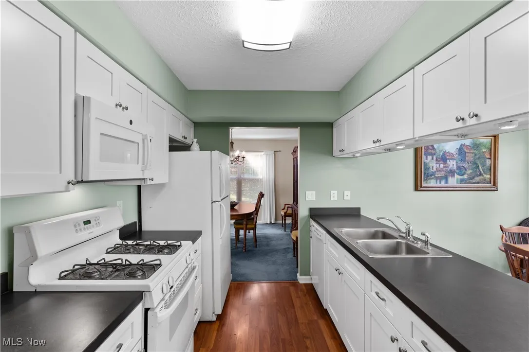 Kitchen featuring white appliances, a textured ceiling, dark countertops, white cabinetry, and dark wood-type flooring