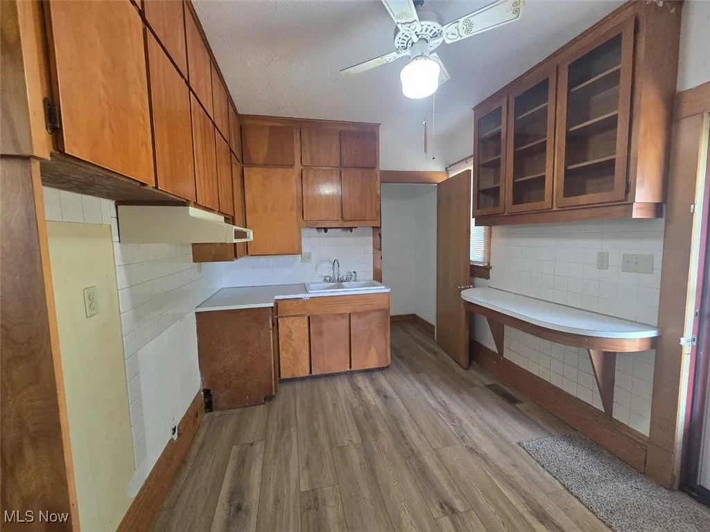 Kitchen featuring brown cabinets, dark wood-type flooring, light countertops, glass insert cabinets, and tasteful backsplash