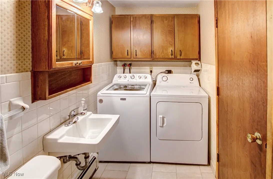 Laundry room inside of bathroom, with tile walls, wallpapered walls, a wainscoted wall, washing machine and dryer, and light tile patterned flooring