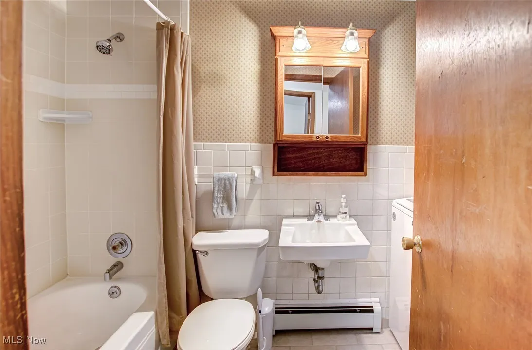 Bathroom featuring shower / tub combo with curtain, tile walls, a baseboard heating unit, a wainscoted wall, and wallpapered walls