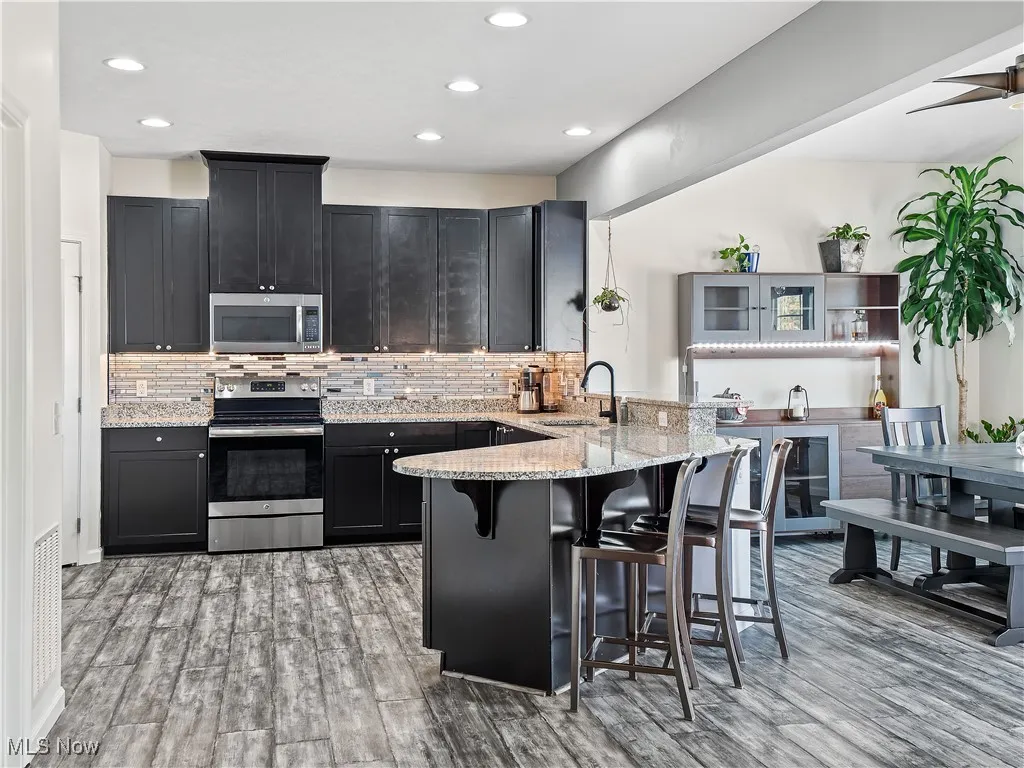 Kitchen featuring dark cabinets, appliances with stainless steel finishes, light stone countertops, a kitchen bar, and a peninsula