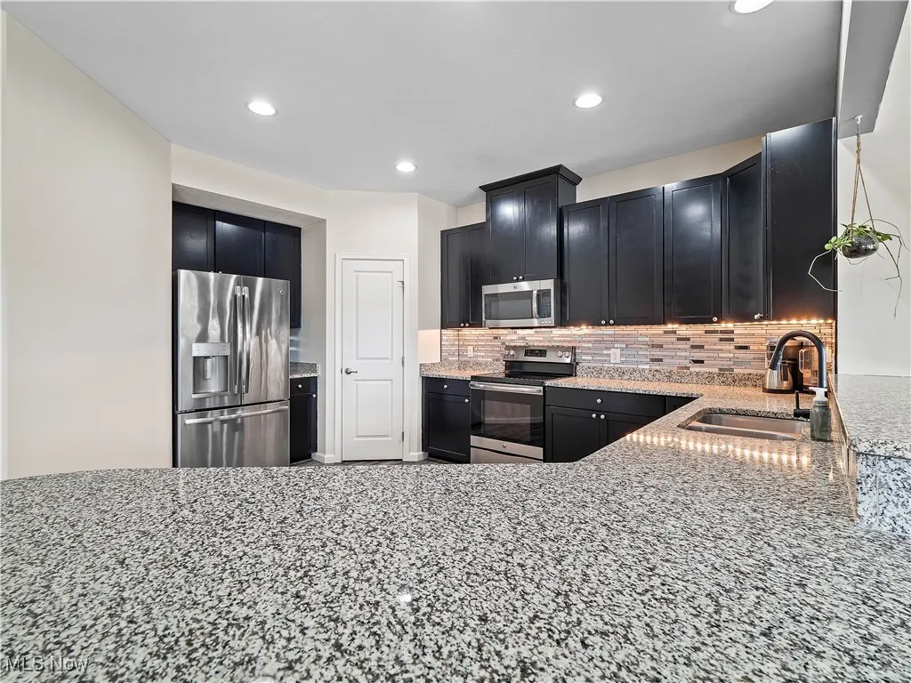 Kitchen with light stone counters, backsplash, dark cabinetry, stainless steel appliances, and recessed lighting