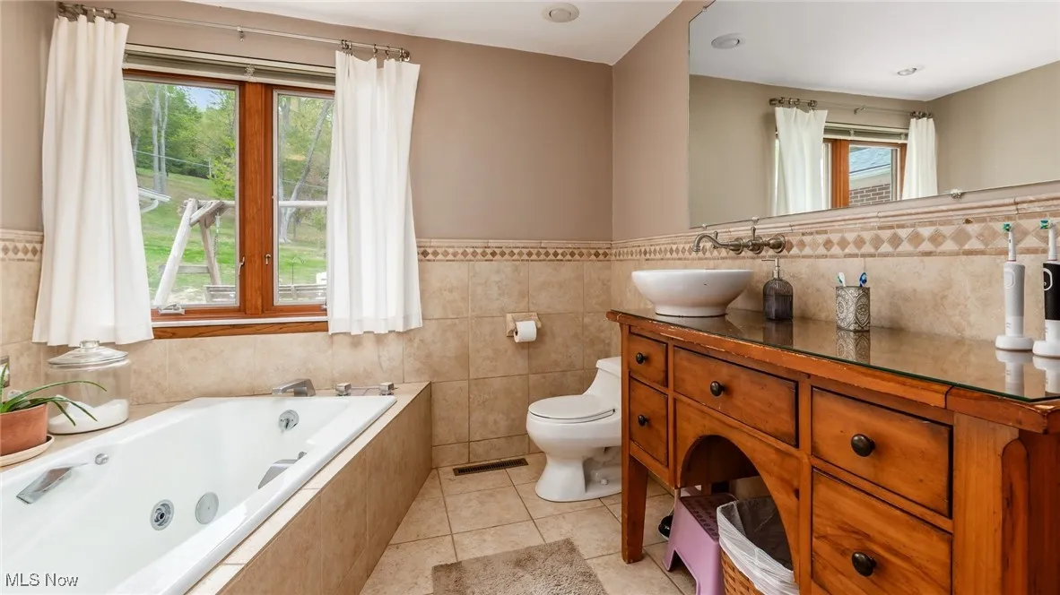 Bathroom featuring tile walls, light tile patterned floors, a whirlpool tub, wainscoting, and double vanity