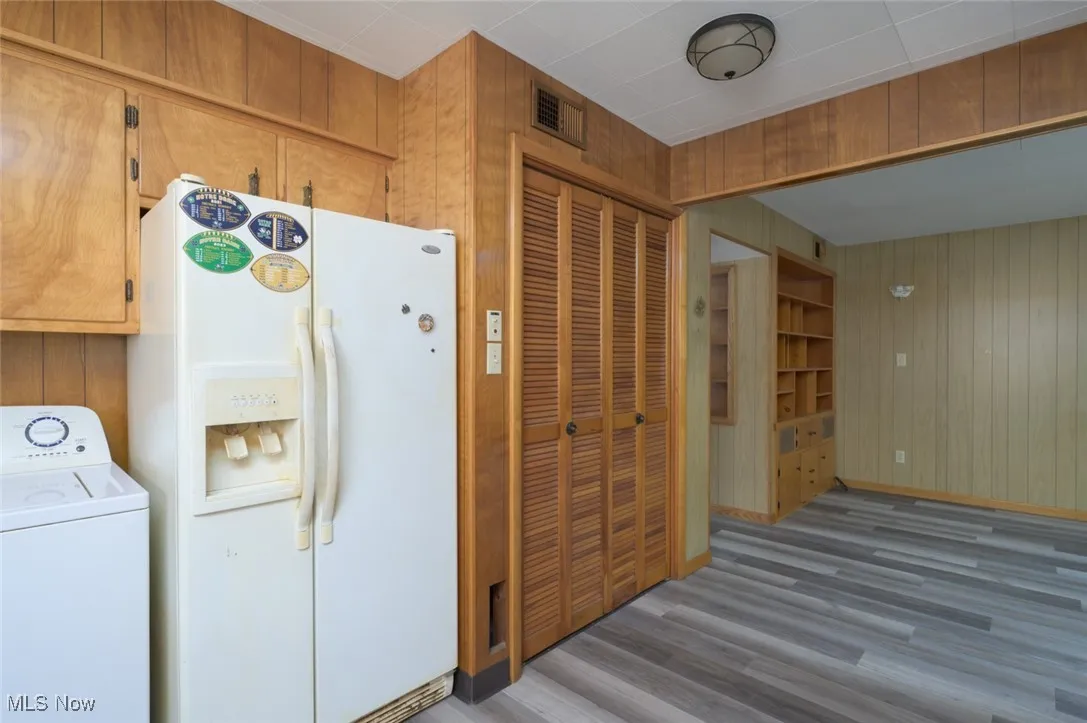 Kitchen with wooden walls, white fridge with ice dispenser, washer / dryer, wood finished floors, and brown cabinets