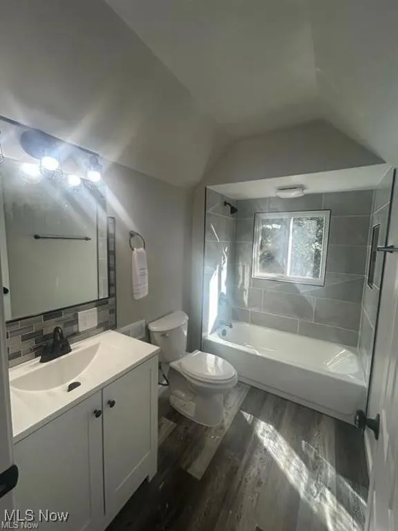 Bathroom featuring vanity, shower / bath combination, vaulted ceiling, dark wood-type flooring, and tasteful backsplash