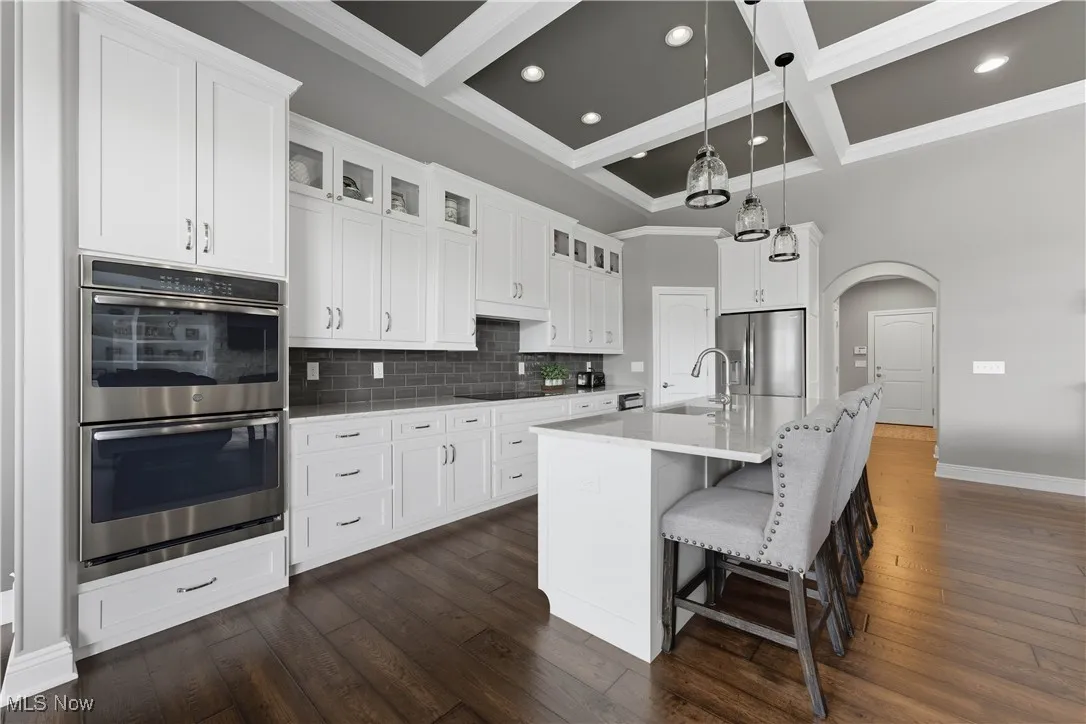 Kitchen featuring glass insert cabinets, arched walkways, decorative backsplash, coffered ceiling, and stainless steel appliances