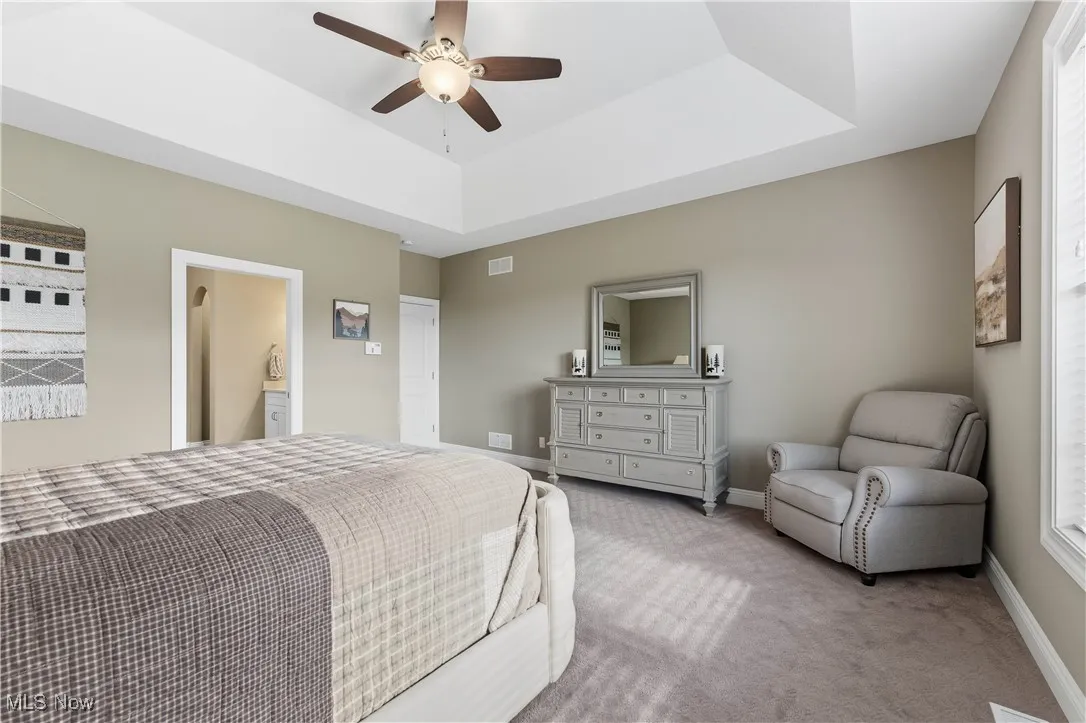 Carpeted bedroom featuring a raised ceiling, a ceiling fan, and ensuite bath