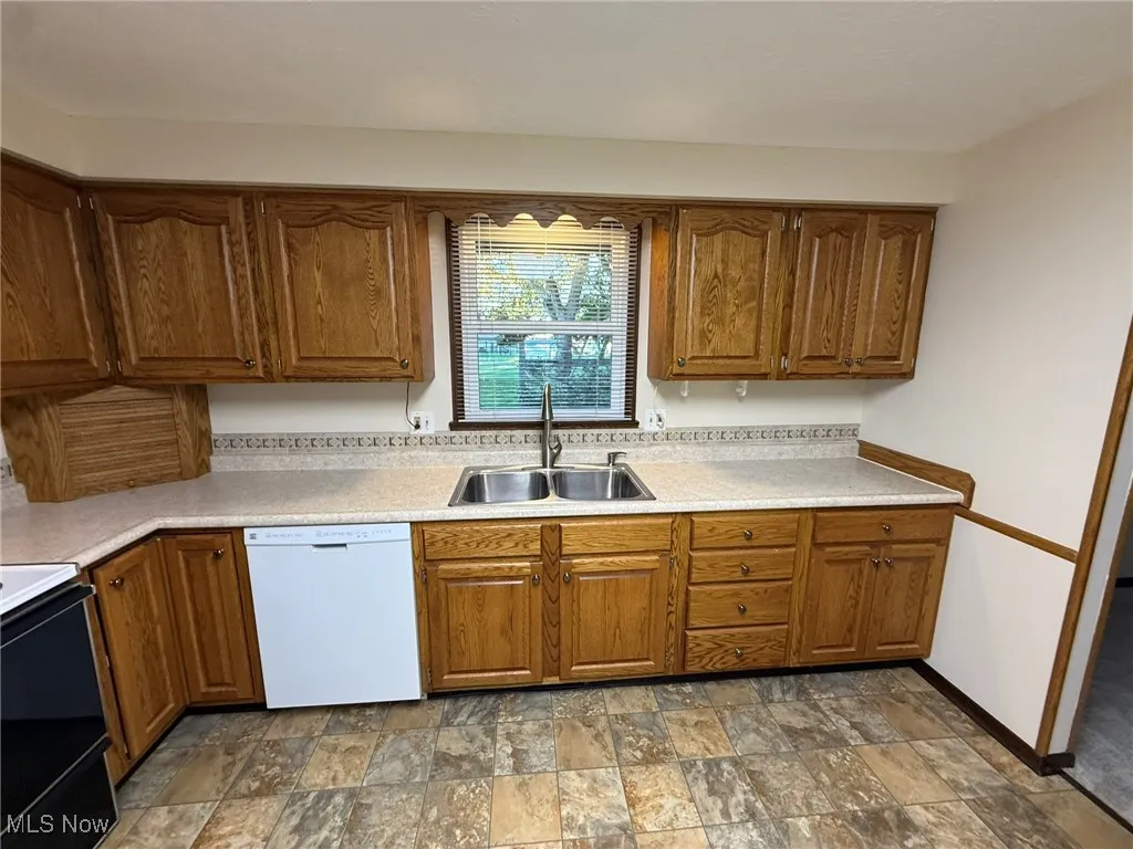 Kitchen with brown cabinets, stone finish floors, white dishwasher, black range oven, and light countertops