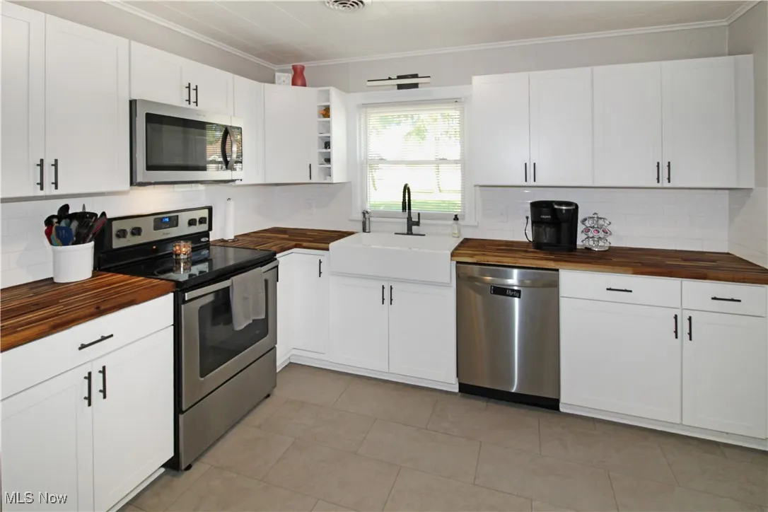 Kitchen with butcher block counters, stainless steel appliances, backsplash, white cabinetry, and crown molding