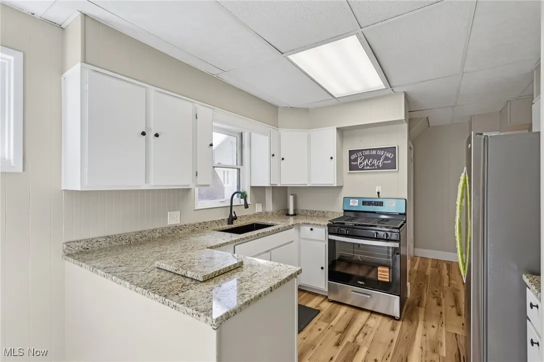 Kitchen with stainless steel appliances, a paneled ceiling, light stone countertops, white cabinetry, and a peninsula