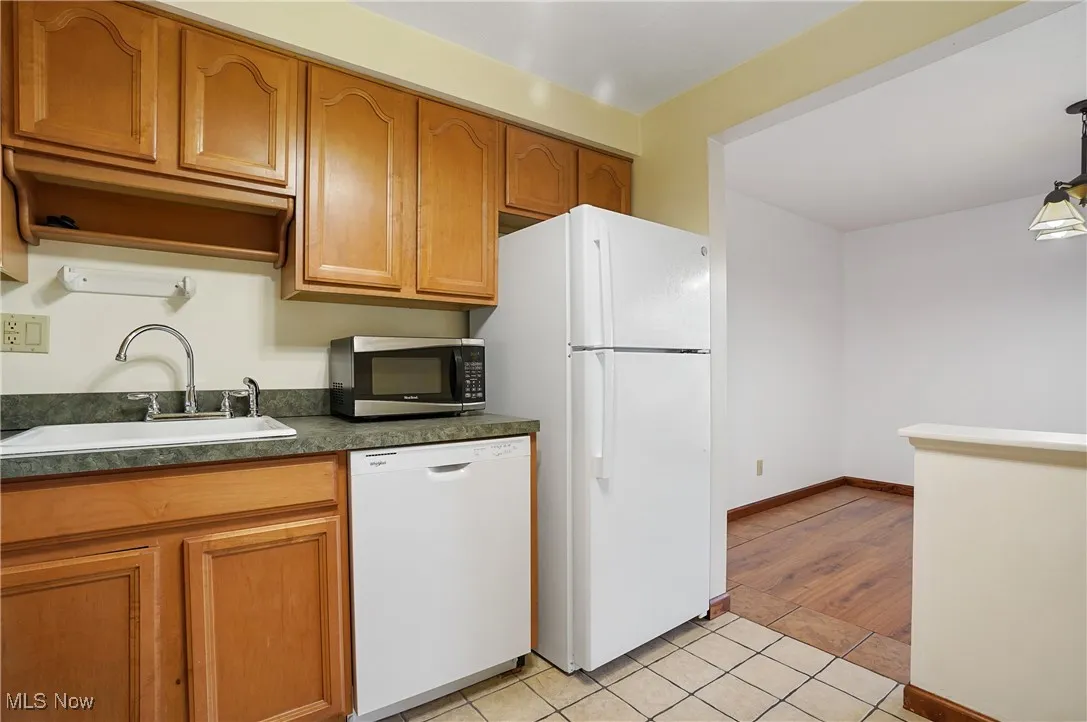 Kitchen with white appliances, light tile patterned floors, brown cabinetry, dark countertops, and hanging light fixtures