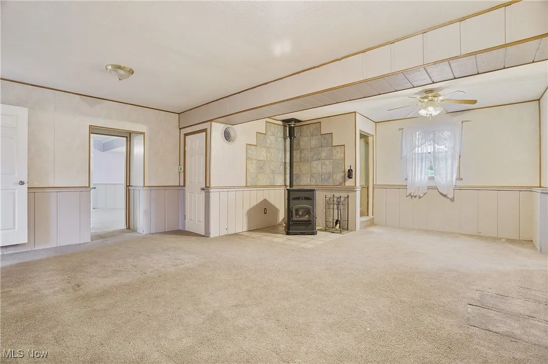 Unfurnished living room with a wood stove, carpet, a ceiling fan, a decorative wall, and a wainscoted wall