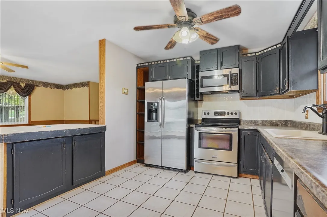 Kitchen featuring ceiling fan, stainless steel appliances, light tile patterned floors, dark countertops, and dark cabinets