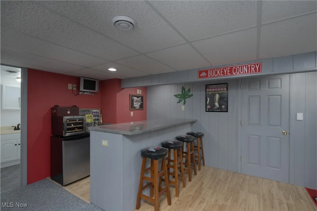 Indoor dry bar with a drop ceiling, stainless steel refrigerator, light wood-type flooring, and wooden walls