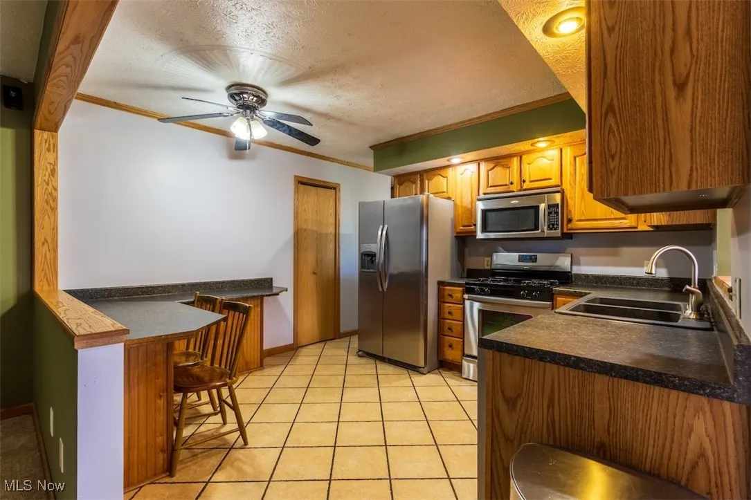 Kitchen featuring ornamental molding, appliances with stainless steel finishes, a textured ceiling, brown cabinetry, and dark countertops