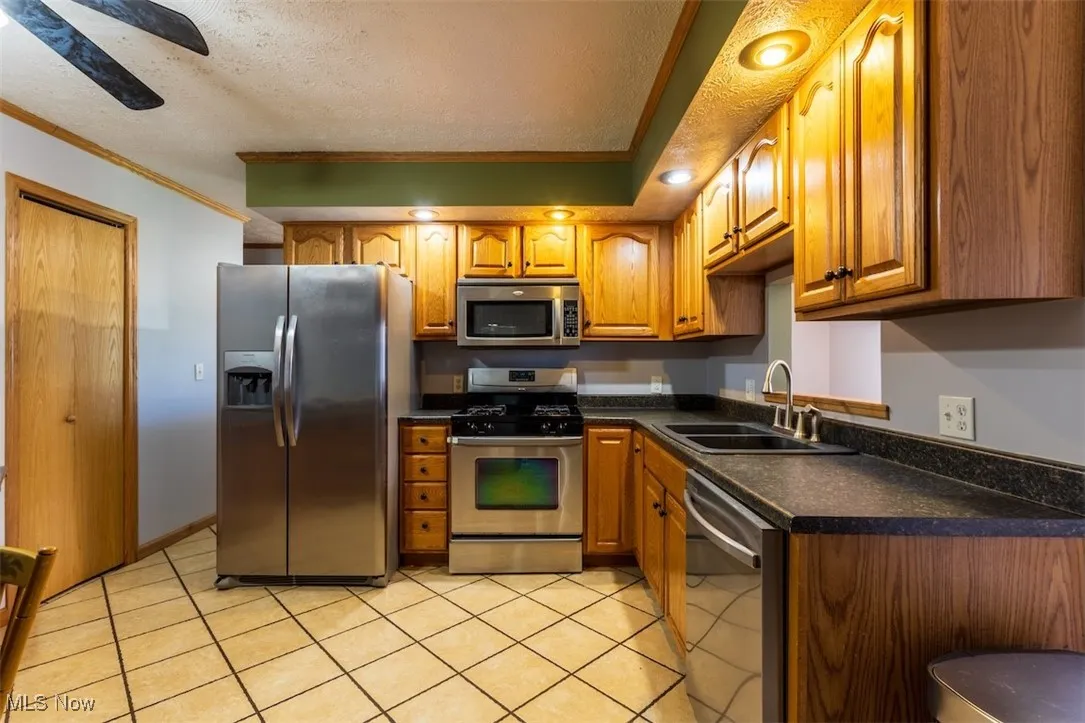 Kitchen featuring brown cabinetry, stainless steel appliances, a textured ceiling, ornamental molding, and dark countertops