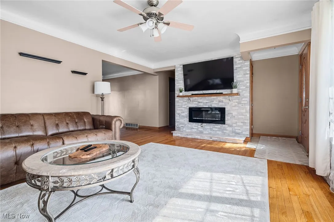 Living room featuring light wood finished floors, a ceiling fan, a stone fireplace, and ornamental molding