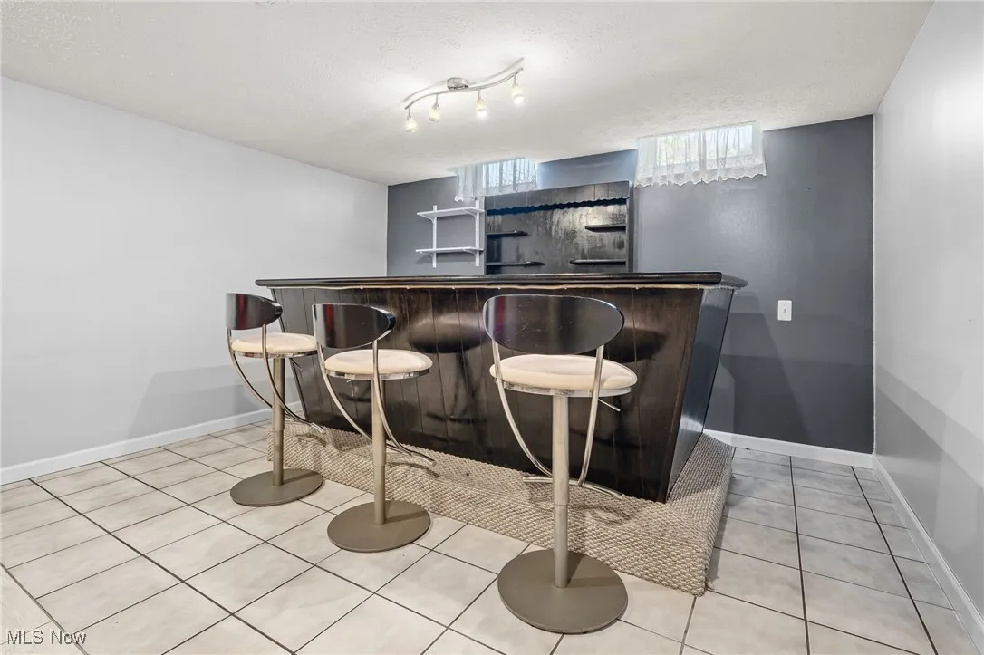 Indoor dry bar featuring a textured ceiling, light tile patterned floors, dark countertops, open shelves, and dark brown cabinetry