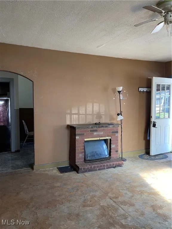 Unfurnished living room with a textured ceiling, a brick fireplace, a ceiling fan, unfinished concrete floors, and arched walkways