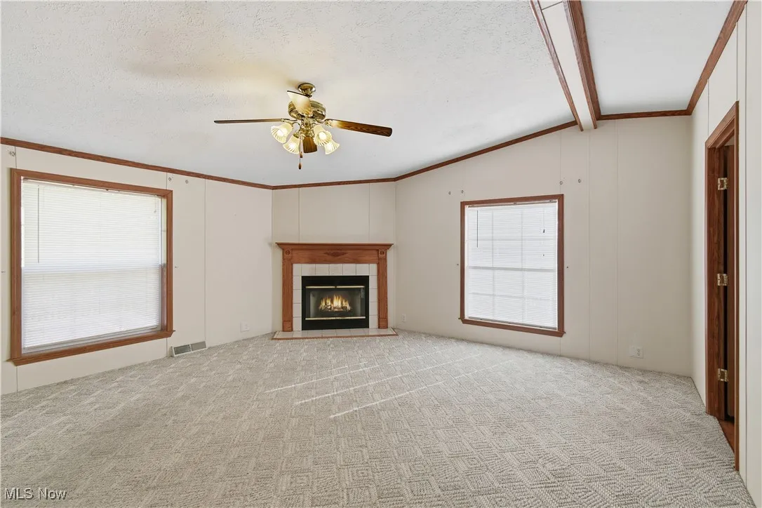 Unfurnished living room with a tile fireplace, ceiling fan, ornamental molding, light carpet, and a textured ceiling