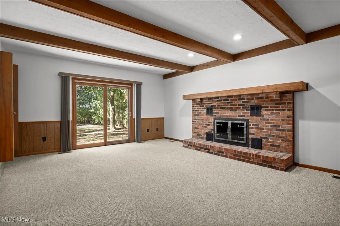 Unfurnished living room featuring a brick fireplace, carpet, beam ceiling, wainscoting, and a textured ceiling
