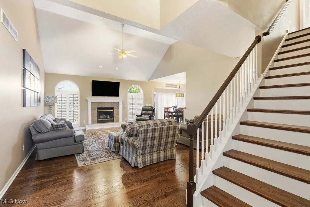 Living area with dark wood-style floors, ceiling fan, high vaulted ceiling, a glass covered fireplace, and stairway