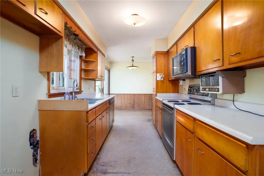 Kitchen with a wainscoted wall, electric stove, brown cabinets, light carpet, and light countertops