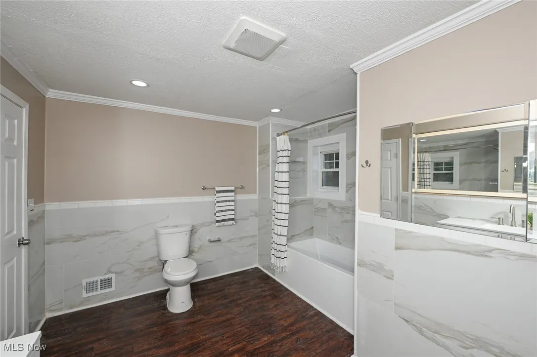 Full bath with tile walls, crown molding, shower / bath combo with shower curtain, a textured ceiling, and wainscoting