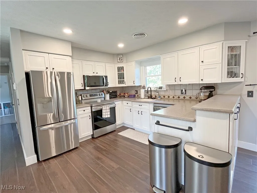 Kitchen with glass insert cabinets, appliances with stainless steel finishes, white cabinets, dark wood-type flooring, and tasteful backsplash