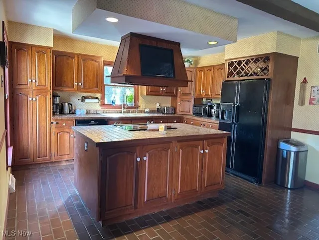 Kitchen with brown cabinets, black refrigerator with ice dispenser, brick patterned flooring, wall chimney range hood, and wallpapered walls