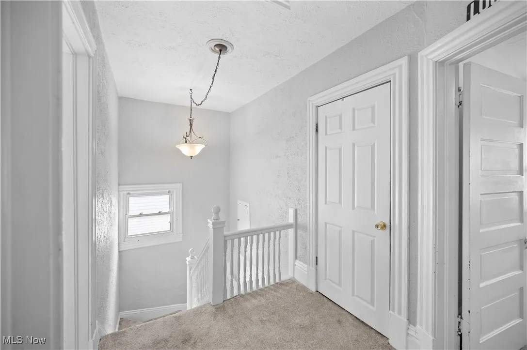 Hallway with an upstairs landing, a textured wall, light colored carpet, and a textured ceiling