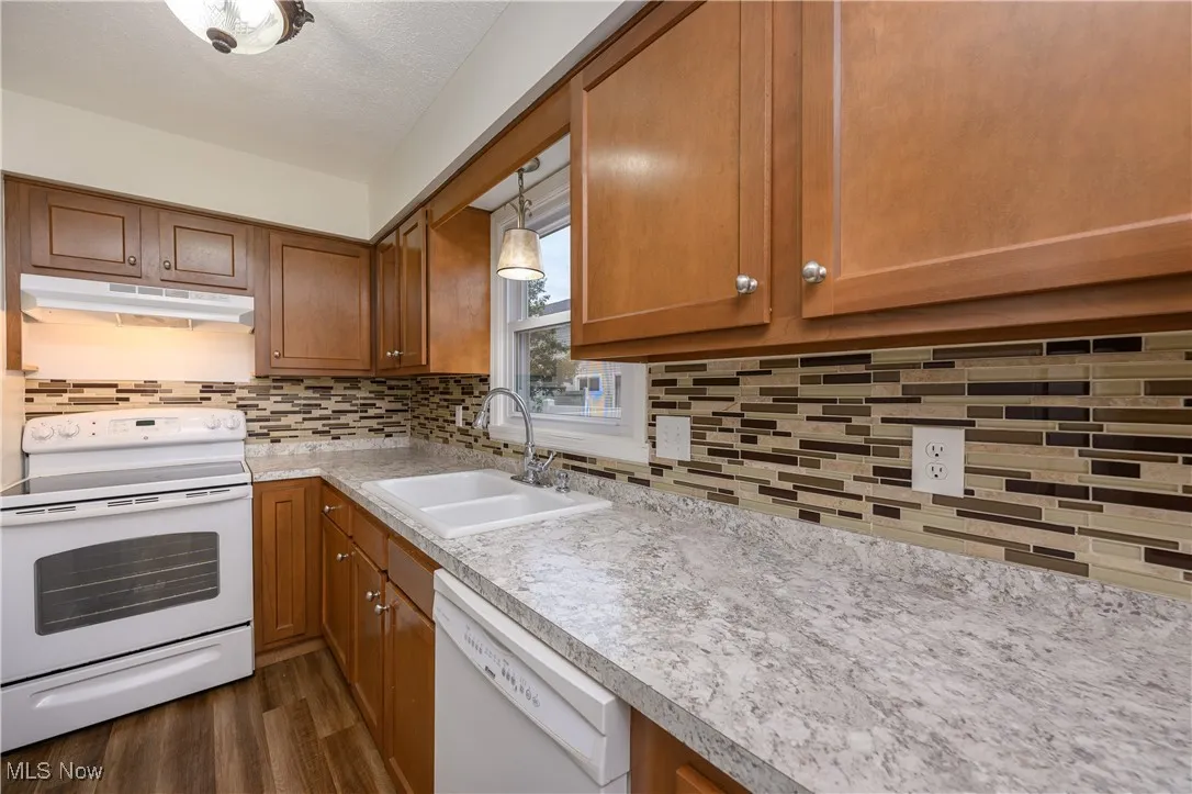 Kitchen featuring white appliances, brown cabinets, light countertops, decorative backsplash, and under cabinet range hood