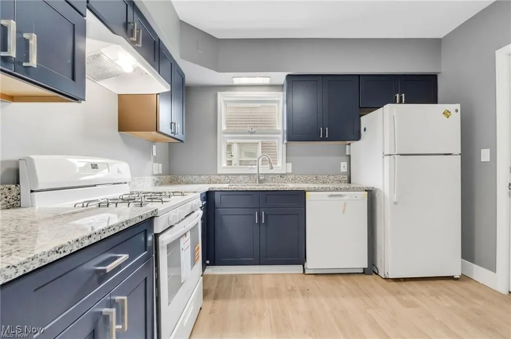 Kitchen featuring white appliances, light wood-style flooring, light stone counters, under cabinet range hood, and blue cabinetry