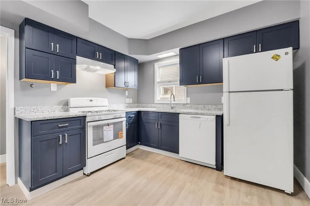 Kitchen with white appliances, blue cabinets, under cabinet range hood, and light wood-type flooring