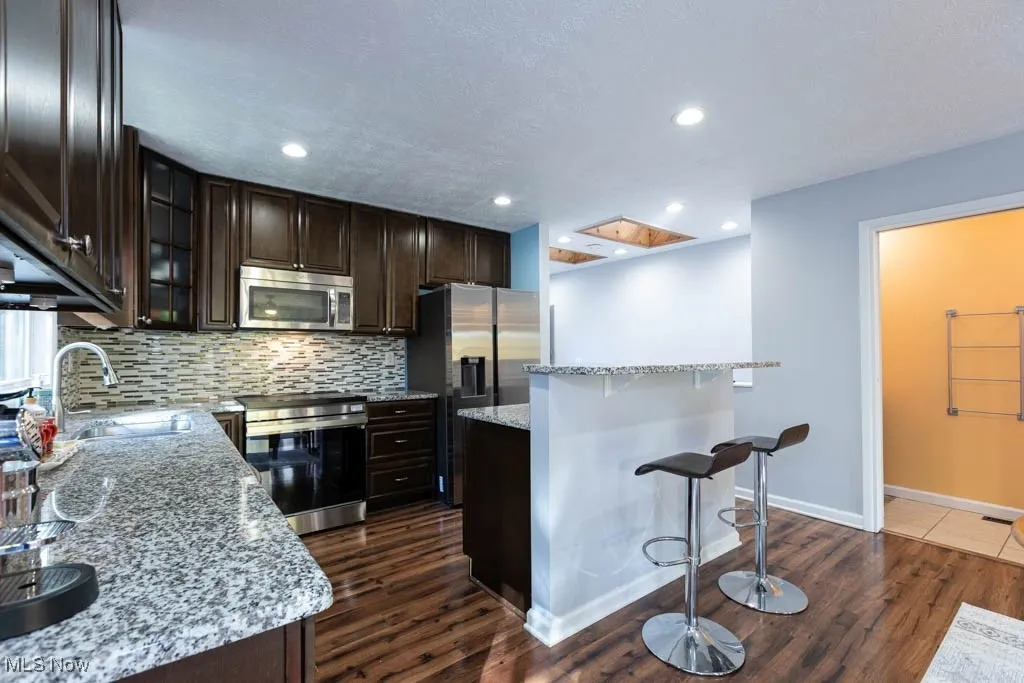 Kitchen featuring dark brown cabinetry, a breakfast bar, glass insert cabinets, appliances with stainless steel finishes, and backsplash