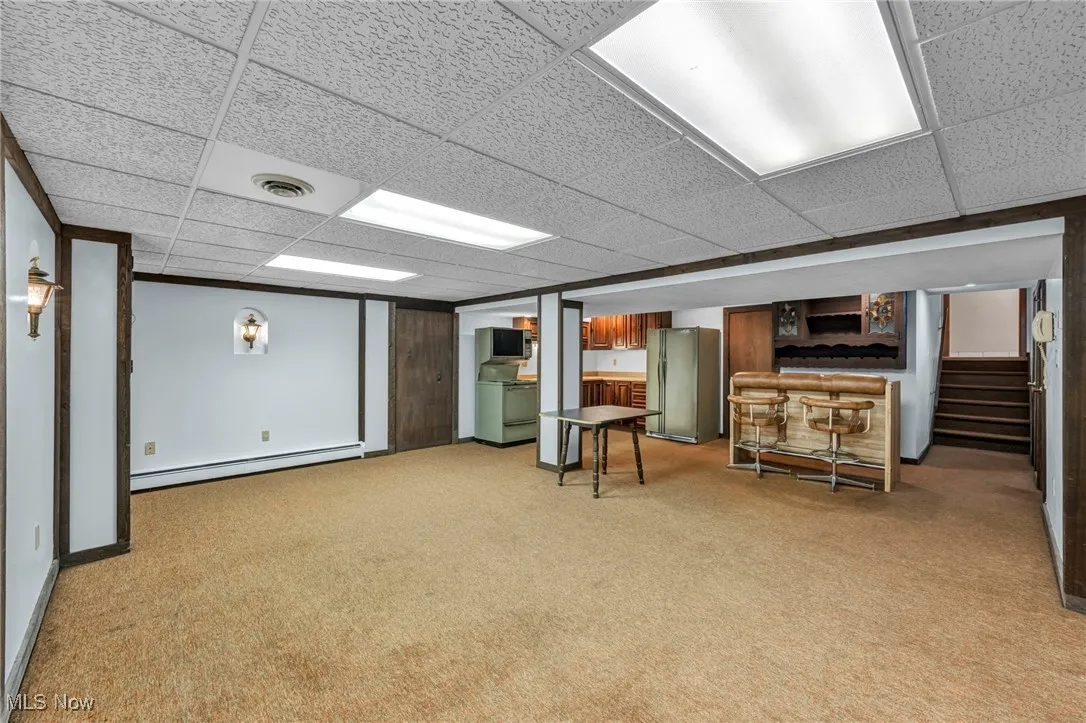 Finished below grade area with light carpet, freestanding refrigerator, a baseboard radiator, stairs, and a paneled ceiling