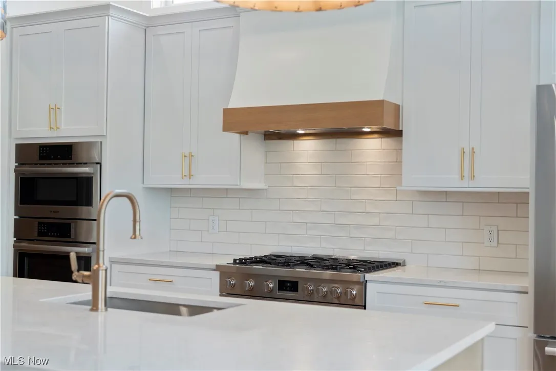 Kitchen with white cabinets, premium range hood, decorative backsplash, light stone counters, and appliances with stainless steel finishes