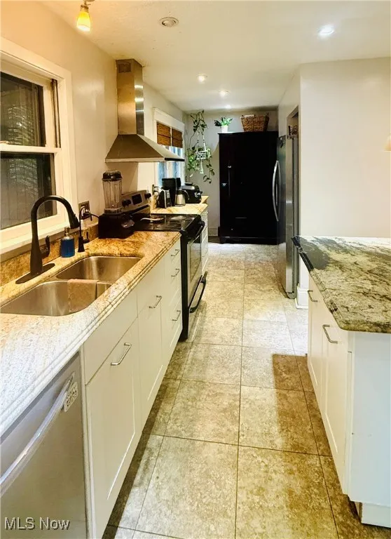 Kitchen with stainless steel appliances, wall chimney exhaust hood, light stone counters, white cabinets, and light tile patterned flooring